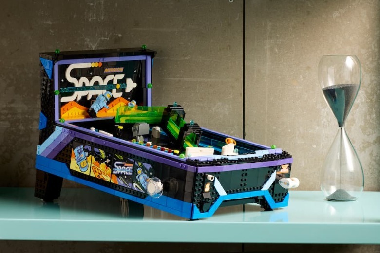 LEGO yapım parçalarıyla inşa edilmiş, üzerinde "Mission Space" yazan retro tarzda bir uzay temalı pinbol (langırt) makinesi modeli. Makine mavi, mor, siyah ve şeffaf yeşil parçalardan oluşuyor ve üzerinde küçük astronot figürleri barındırıyor. Parlak, açık mavi bir rafın üzerinde duran bu modelin hemen sağında, içinde koyu renkli kum olan cam bir kum saati yer alıyor. Arka planda ise pürüzsüz gri bir duvar bulunuyor.